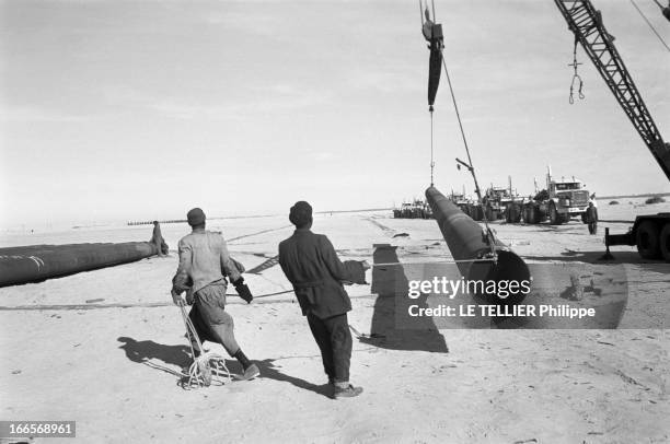 Construction Of Pipe Line. En 1958, dans le désert du Sahara, deux ouvriers manoeuvrant un segments de canalisation suspendu à un treuil au moyen...