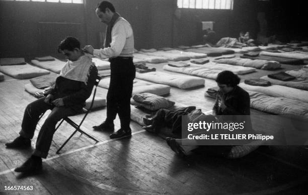 The Action Of Abbe Pierre. Ile-de-France- Février 1954- L'Association Emmaüs: dans un gymnase, un compagnon coiffe un homme assis sur un siège,...