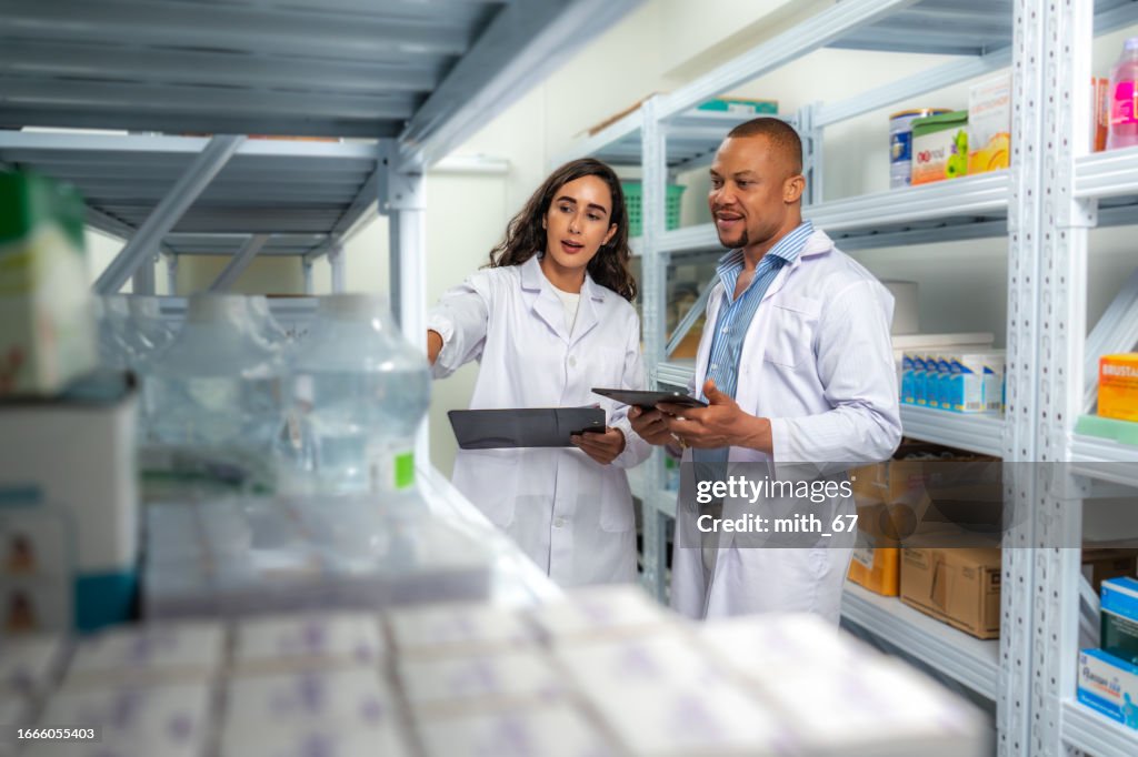 Beautiful Latin pharmacist woman looking at medicine list from her record document for checking medicine stock with her African American male coworker at drug store inventory room. Pharmacist black man and tablet for medicine management