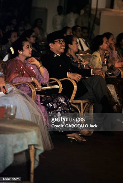 Sukarno, President Of Indonesia. A Djakarta, parmi un public, assis au premier rang dans un siège en rotin, encadré de femmes non identifiées, le...