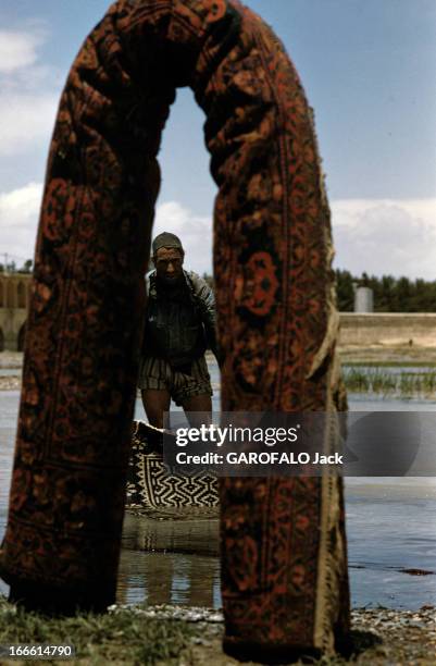 The Iranian Rugs. En Iran, à Ispahan, un homme baigne un tapis dans l'eau du Zâyandé-Roud, derrière un tapis enroulé, posé debout en forme d'arc.