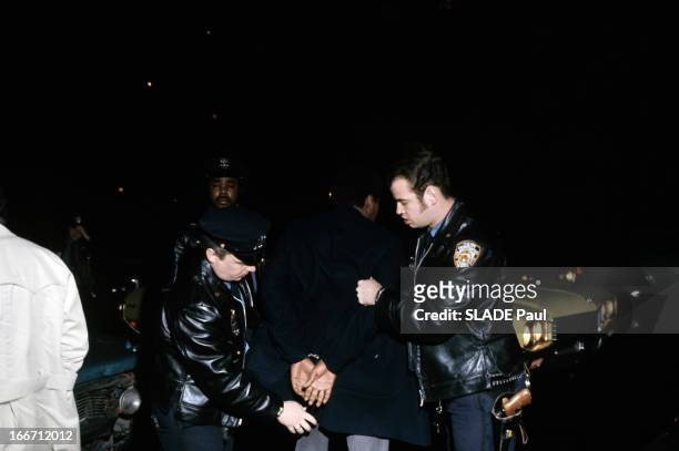 New York, World Capital Of Violence. A Brooklyn, dans une rue, deux policiers, en blouson de cuir avec une casquette, menottant un homme noir.