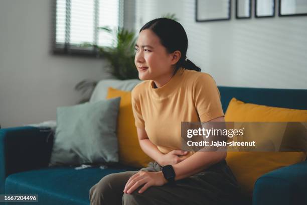 asian woman feeling stomachache sitting on sofa in living room at home. - fart stock pictures, royalty-free photos & images