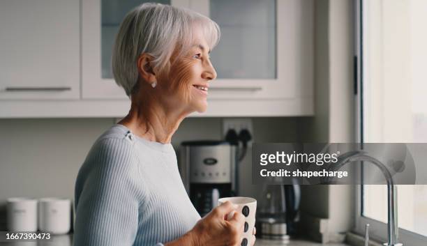 caffè, sorriso e una donna anziana che pensa nella cucina della sua casa mentre sogna ad occhi aperti al mattino. tè, idea e un anziano felice che guarda attraverso la finestra mentre beve una bevanda calda - woman smile kitchen foto e immagini stock