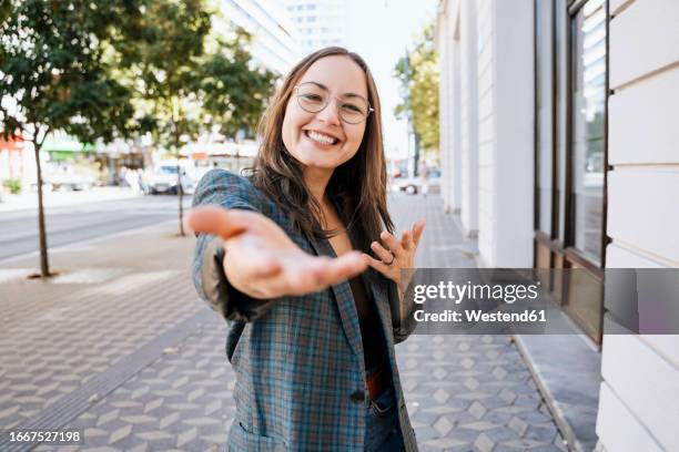 smiling woman gesturing near building at footpath - inviting gesture stockfoto's en -beelden
