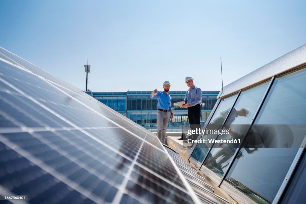 Two businessmen wearing hardhats having a meeting on the roof of a company building with solar panels