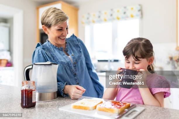 girl drinking from mug by mother in kitchen at home - girl making sandwich stock pictures, royalty-free photos & images