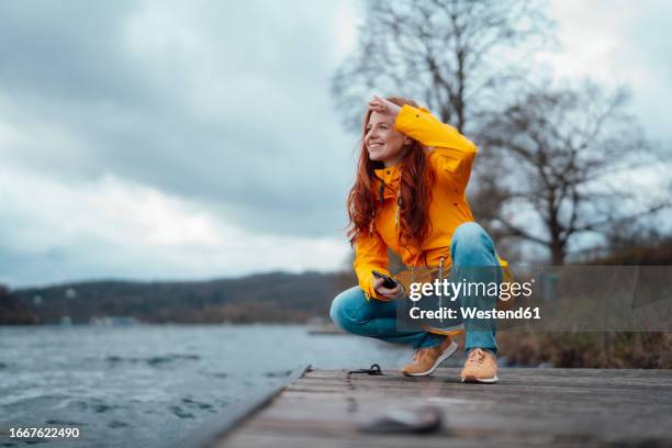 smiling woman squatting on pier by lake - レインコート ストックフォトと画像