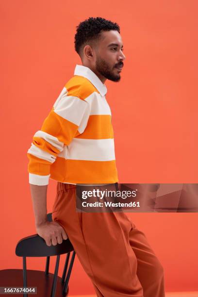 young man leaning on chair against orange background - man leaning chair standing side view stock pictures, royalty-free photos & images