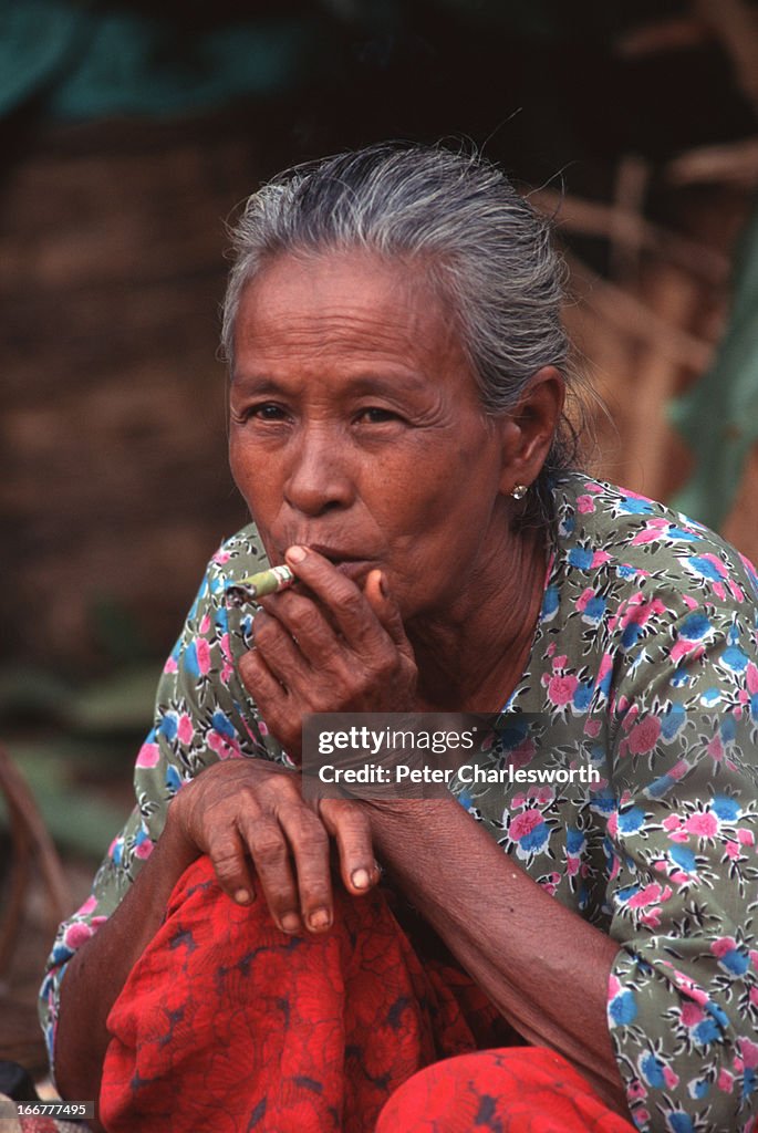 An older woman, a market vendor, takes a break to smoke a...