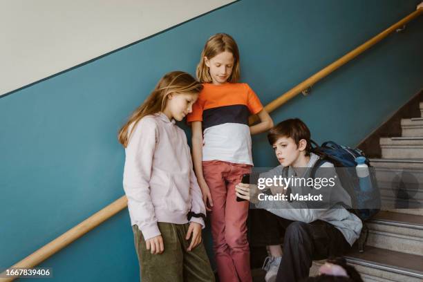 boy sharing smart phone with female friend standing against wall on school building - schulkind stock-fotos und bilder