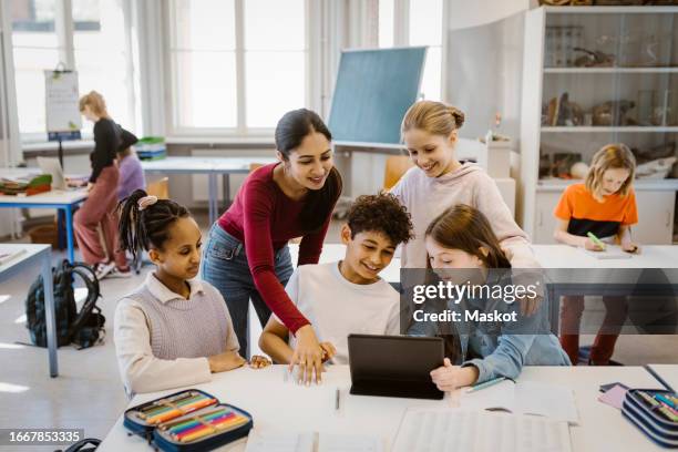 teacher with male and female students using tablet pc at desk in classroom - human age stock pictures, royalty-free photos & images