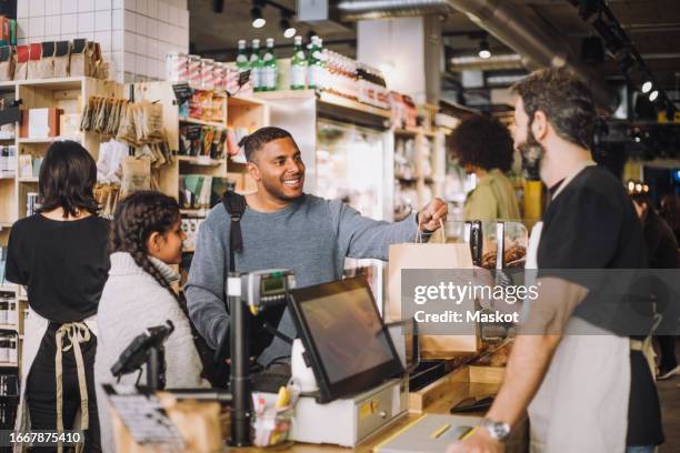 male retail clerk giving shopping bag to customers at checkout in grocery store - arab shopping stockfoto's en -beelden