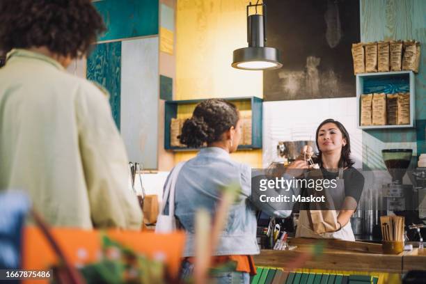 saleswoman giving shopping bag to female customer while man waiting in line at checkout - supermarket queue stock pictures, royalty-free photos & images