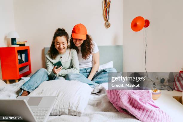 happy young woman showing smart phone to female friend while spending leisure time at home - pillow icon stock pictures, royalty-free photos & images