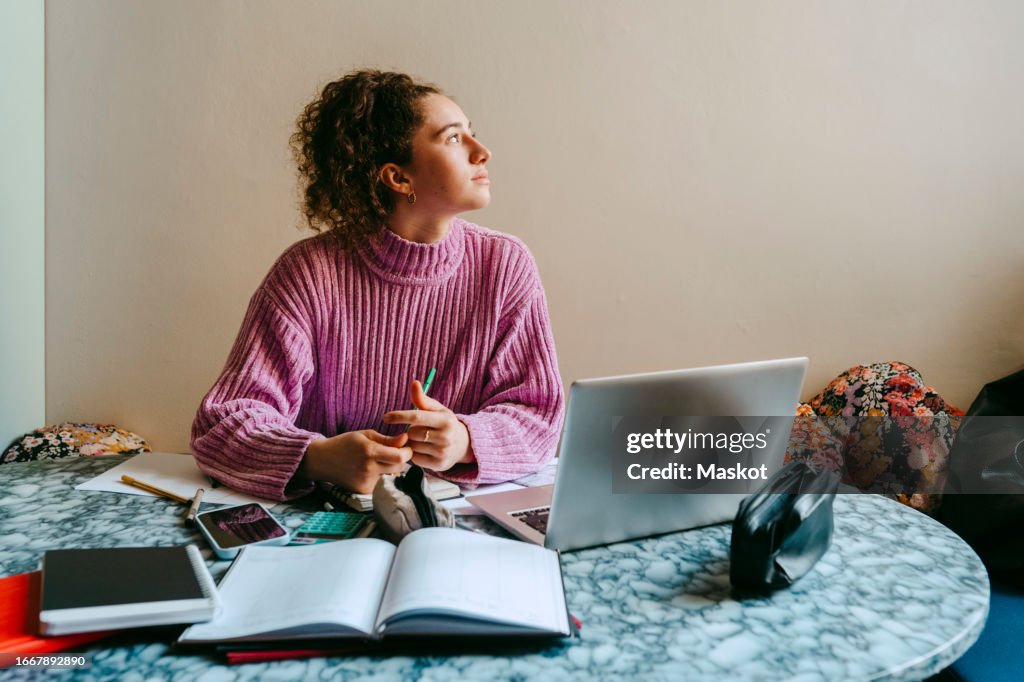Thoughtful young woman looking away while sitting with laptop and books at table