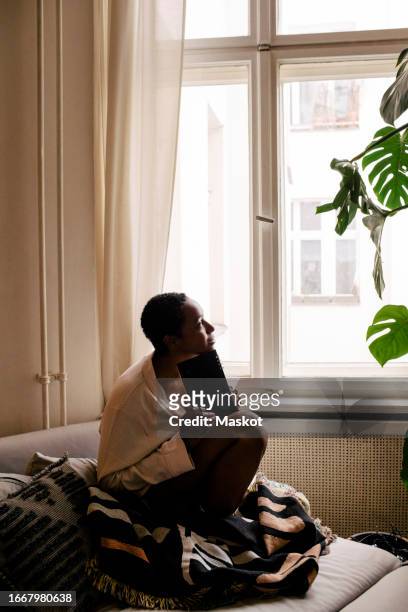 thoughtful young woman with book sitting on bed at home - literatur stock-fotos und bilder