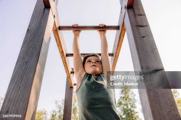 low angle view of determined boy hanging while doing monkey bars at summer camp - monkey bars fotografías e imágenes de stock