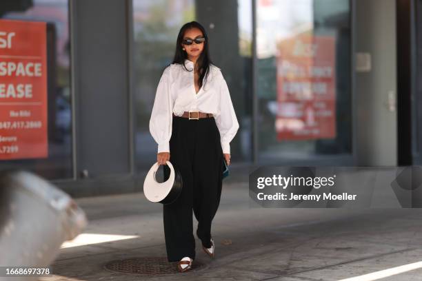Fashion Week guest is seen wearing black sunglasses, golden earrings, a white unbuttoned shirt with sewed-on flowers and puffy long sleeves, a brown...