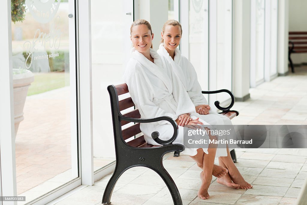 Women in bathrobes sitting on bench poolside at spa