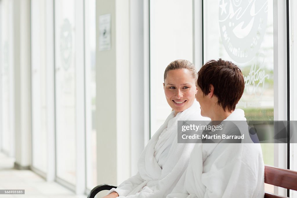 Women in bathrobes sitting on bench poolside at spa