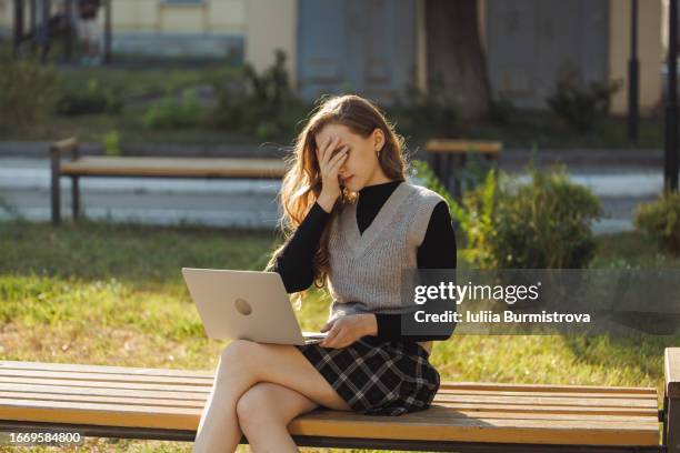 elegant young lady sitting on bench with laptop on knees covering face with hand being upset - sleeveless sweater stock pictures, royalty-free photos & images