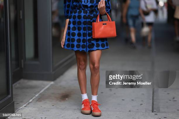 Fashion Week guest is seen wearing big sunglasses with orange lenses, a blue mini dress with a pattern of black polka dots from Kate Spade, golden...
