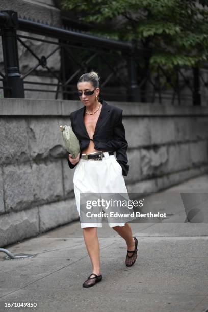 Guest wears sunglasses, a black oversized blazer jacket, a belt, white shorts, mesh shoes, a green bag, outside Khaite , during New York Fashion...