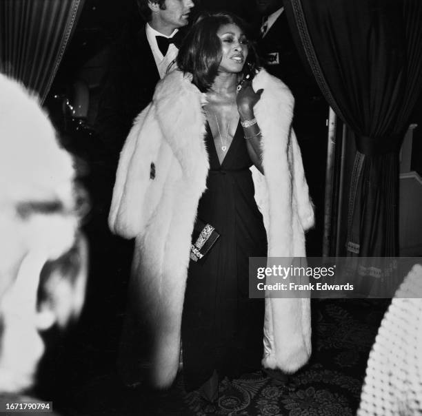 American singer and actress Tina Turner, with a white fur coat draped over her shoulders, over a black dress with a plunging neckline, attends an...