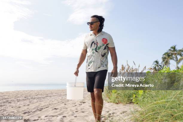 a multiracial millennial man with dreadlocks walks along the beach with a white bucket looking for garbage while wearing casual clothing. - three quarter length stock pictures, royalty-free photos & images
