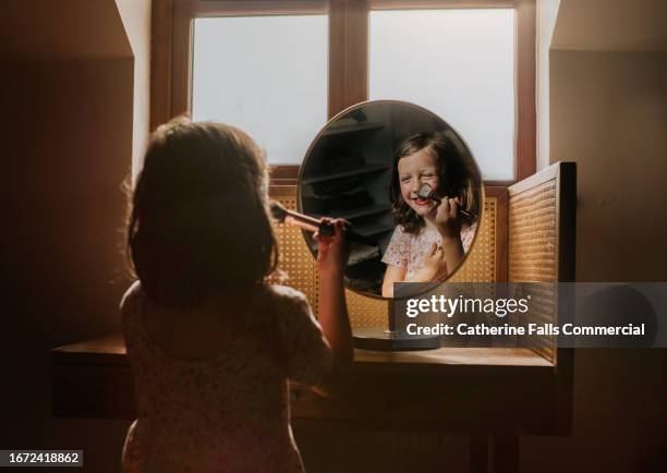 a mischievous little girl applies blusher in a round mirror at a vanity table - kaptafel stockfoto's en -beelden