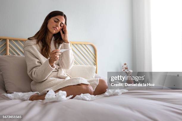 young caucasian woman checking temperature with thermometer, sitting in bed feeling unwell. woman sick in bed. - thermomètre à affichage digital photos et images de collection