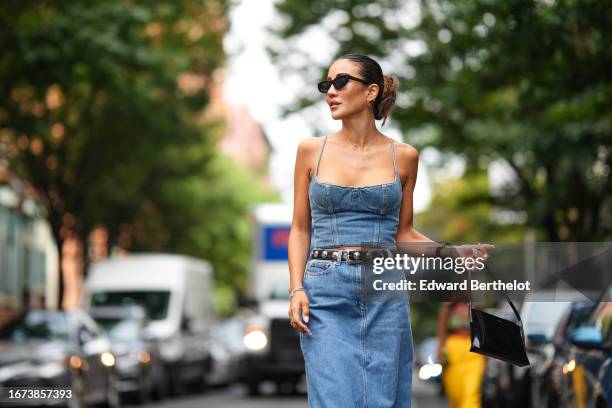 Tamara Kalinic wears sunglasses, a blue denim off-shoulder low neck cropped top, a black leather studded belt, a blue denim maxi skirt, a black...