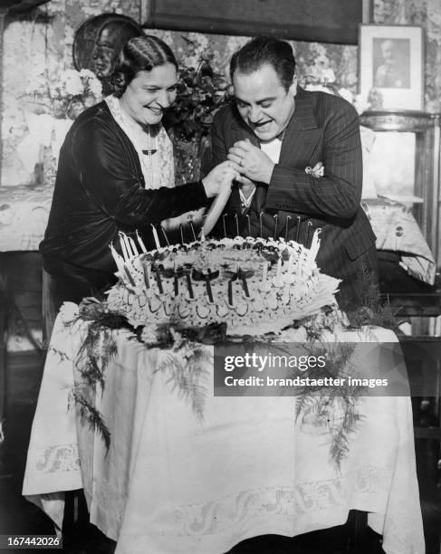 Italian tenor Beniamino Gigli celebrates his 38th Birthday with his wife. The couple cut the cake together. New York. Photograph. 1928. Der...