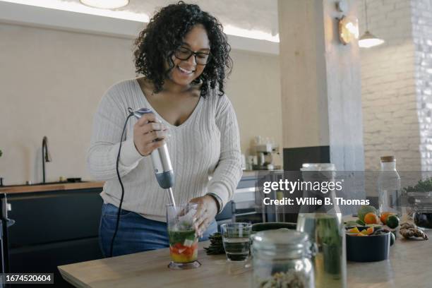 woman using hand blender to make smoothie in kitchen - blender stock-fotos und bilder