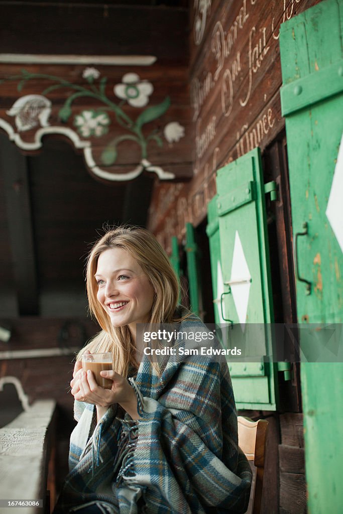 Smiling woman wrapped in a blanket and drinking coffee on cabin porch