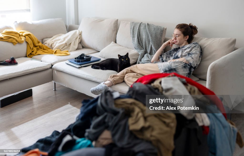 Ein Haufen Kleidung, die nach der Wäsche verstreut liegt und darauf wartet, sortiert zu werden. Faule junge Frau, die eine Pause mit schwarzer Katze auf Sofa auf Hintergrund macht
