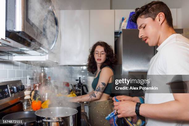 latino man with mustache help his wife to cook, opening bag of potatoes in kitchen. brunette woman in eyeglasses giving cooking instructions to boyfriend in steamy kitchen - gekookte aardappel stockfoto's en -beelden