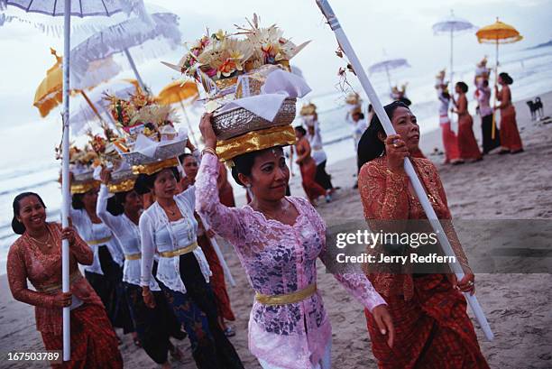 Women tote offering baskets of food, flowers and drink to the beach during a Hindu ceremony. Brightly-dressed women with burgeoning baskets perched...