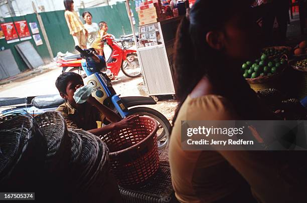 Along crouches behind piles of produce at the Old Market. He is sniffing glue from a plastic bag as he watches a movie playing on a TV in an open-air...