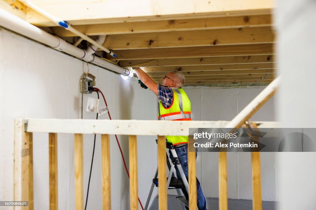 電気ボックスの近くで水漏れの危険があります。地下室の損傷後に壊れた水道管を検査するベストを着た成熟した白髪の労働者