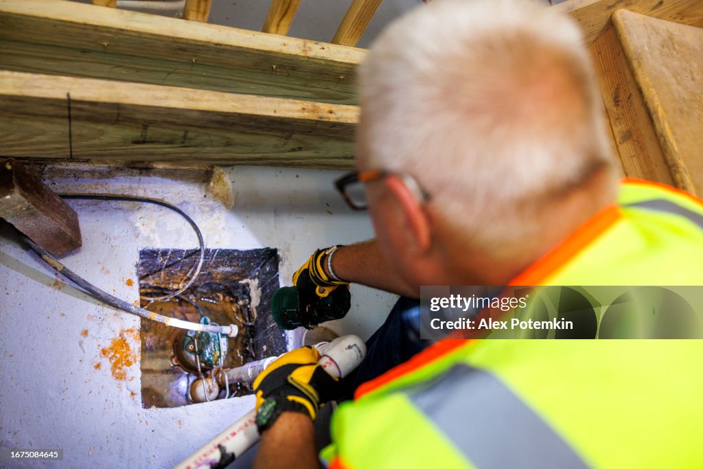 Inspektion des Schadenagenten der Sumpfgrube. Reifer grauhaariger Schadenagent in Weste, der das Entwässerungssystem im Keller inspiziert. Konzentrieren Sie sich in der obigen Ansicht auf die Sumpfpumpe