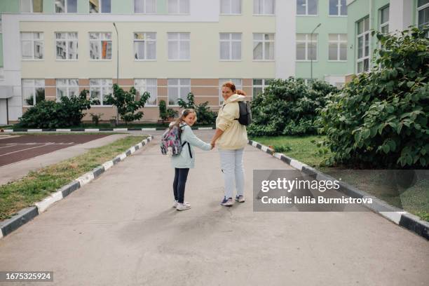 pretty little schoolgirl walking to school hand-in-hand with happy mother, both taking moment to pause and look back at camera - public school building stock pictures, royalty-free photos & images