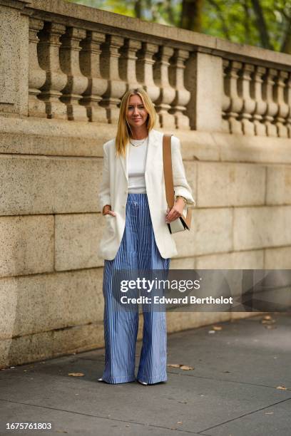 Lisa Aiken wears a necklace, a white oversize blazer jacket, a blue and white striped wide-leg flare pants, a brown and white leather bag, outside...