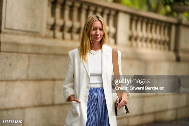 Lisa Aiken wears a necklace, a white oversize blazer jacket, a blue and white striped wide-leg flare pants, a brown and white leather bag, outside...