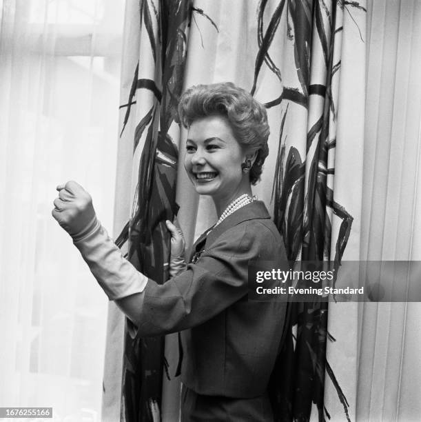 American actress Mitzi Gaynor smiles while clenching a fist beside a curtain, April 24th, 1958. Gaynor was in London for the premiere of the film...