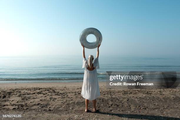 a young woman holds up an inflatable swim ring by the beach - null stock-fotos und bilder