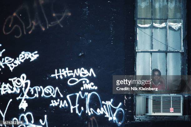 View of a woman, her hand on her cheek, as she looks out of the screen-covered window of a Lower East Side building, New York, New York, January...