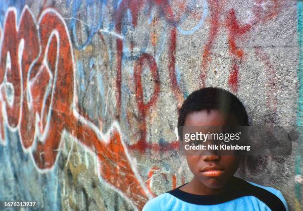 Portrait of an unidentified boy as he leans against a schoolyard wall, which is spray-painted with graffiti, in Cambria Heights, Queens, New York,...
