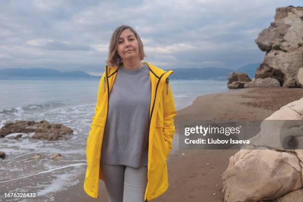 portrait of mature woman (40-45 years) in yellow raincoat spending time on empty beach and looking at camera on cloudy day. - 40 44 years woman caucasian stockfoto's en -beelden